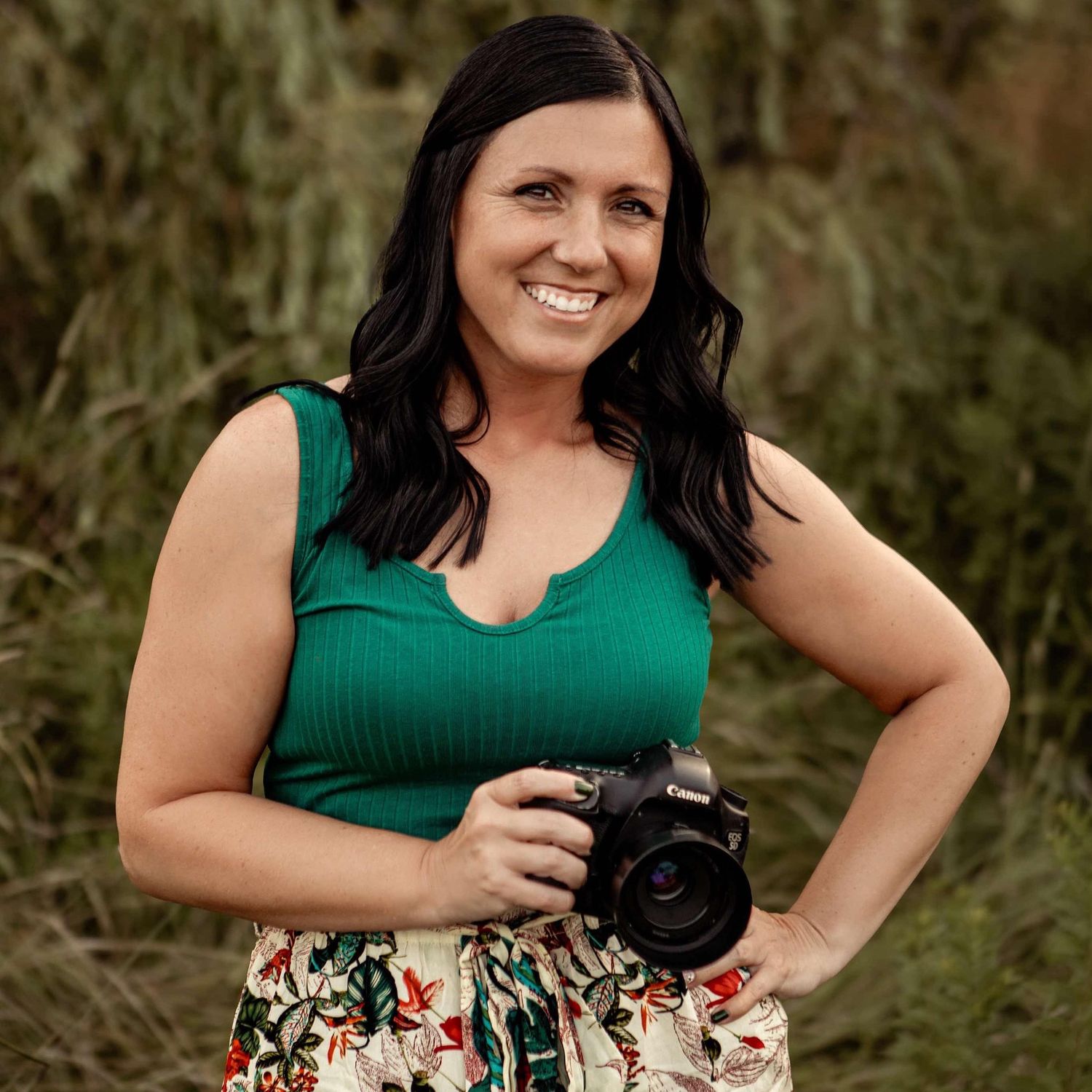 Woman in green top and floral skirt holding a professional camera during an outdoor photoshoot.