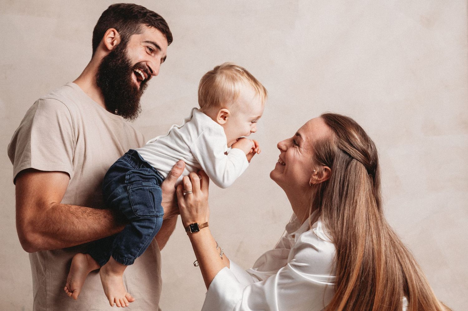 Parents hold and play with their baby in a series of candid lifestyle photos against a neutral wall background.