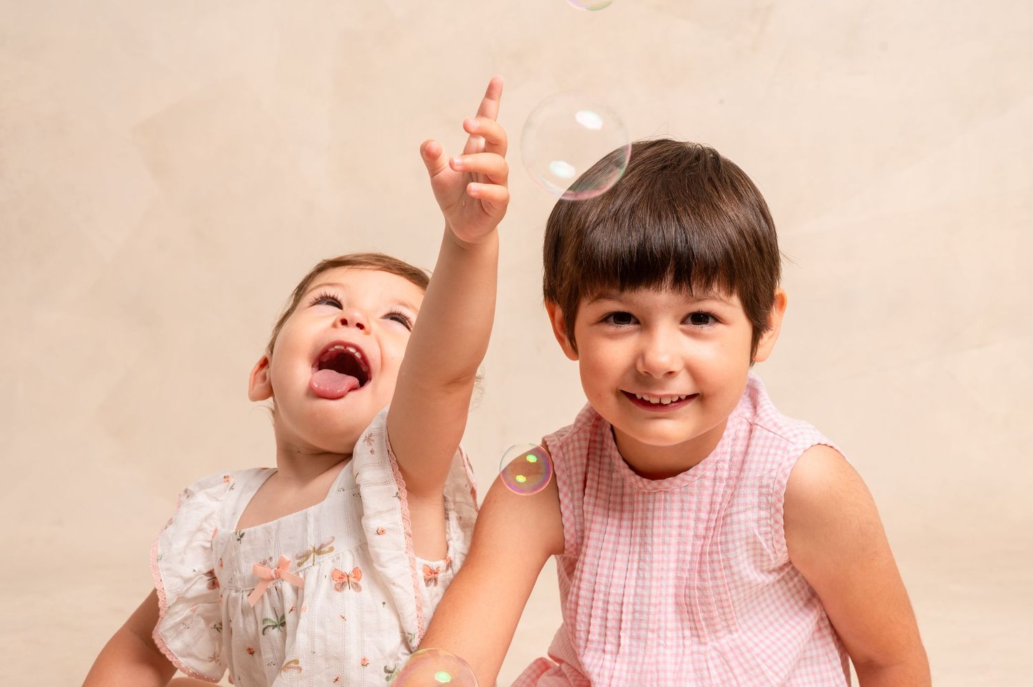 Playful children laugh and point upwards while sitting together against a beige backdrop.