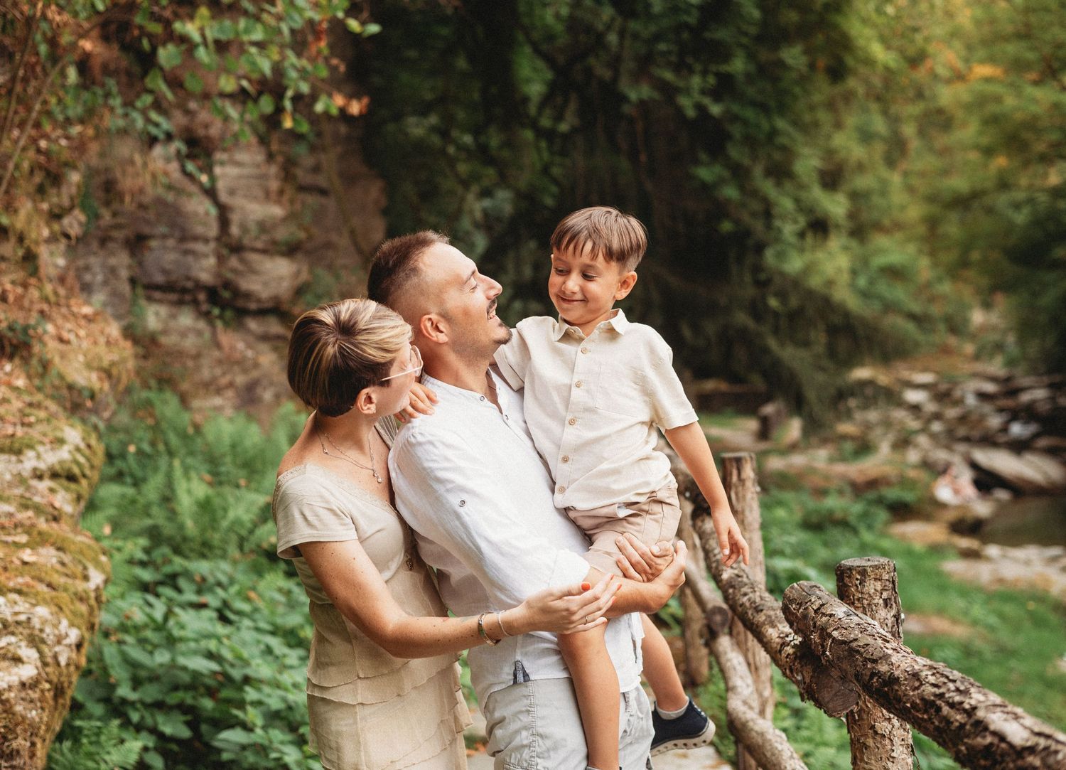 Family of three poses together outdoors in white clothing near a creek and rock wall on a sunny day.