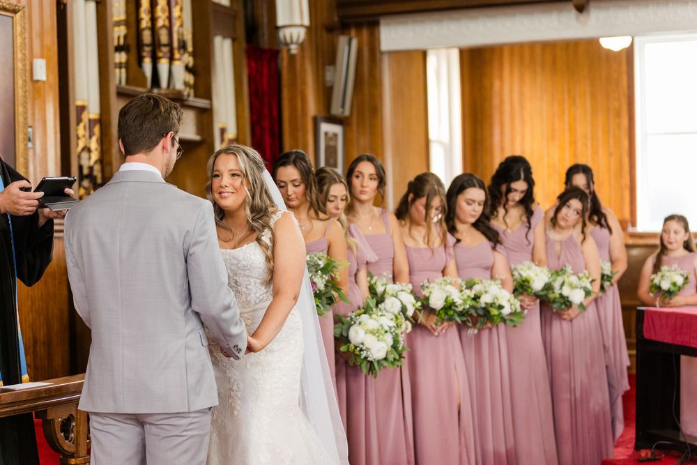 Wedding party stands at altar with bridesmaids wearing dusty rose gowns and holding white floral bouquets at ceremony.