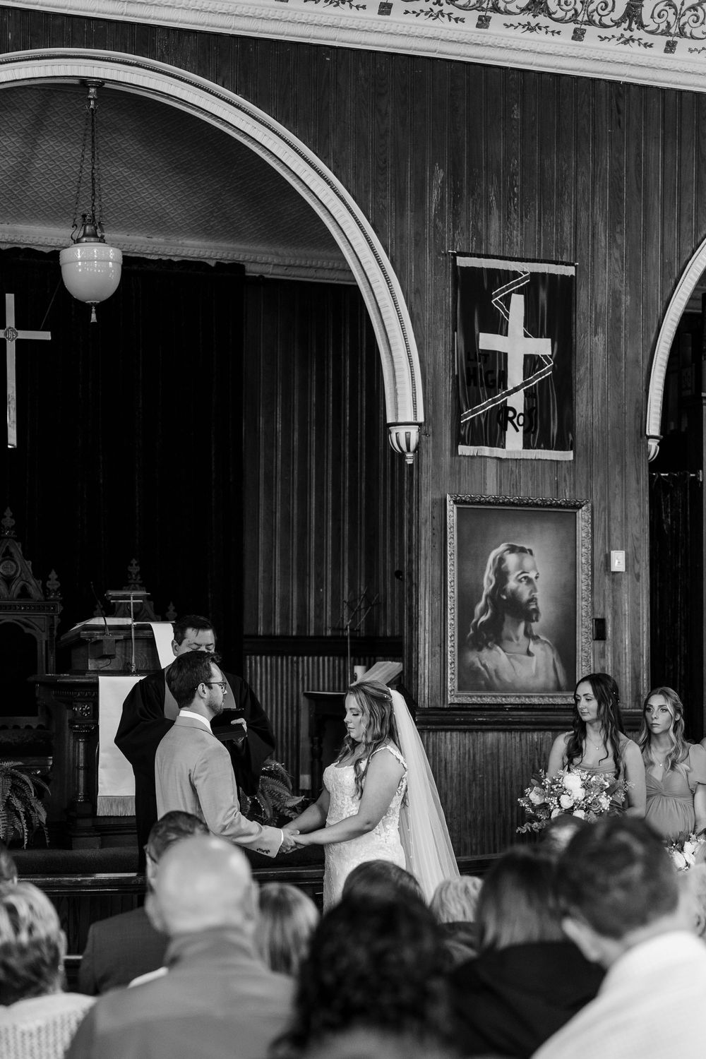 A wedding ceremony in progress with guests seated in rows watching the couple at an indoor venue with arched details.