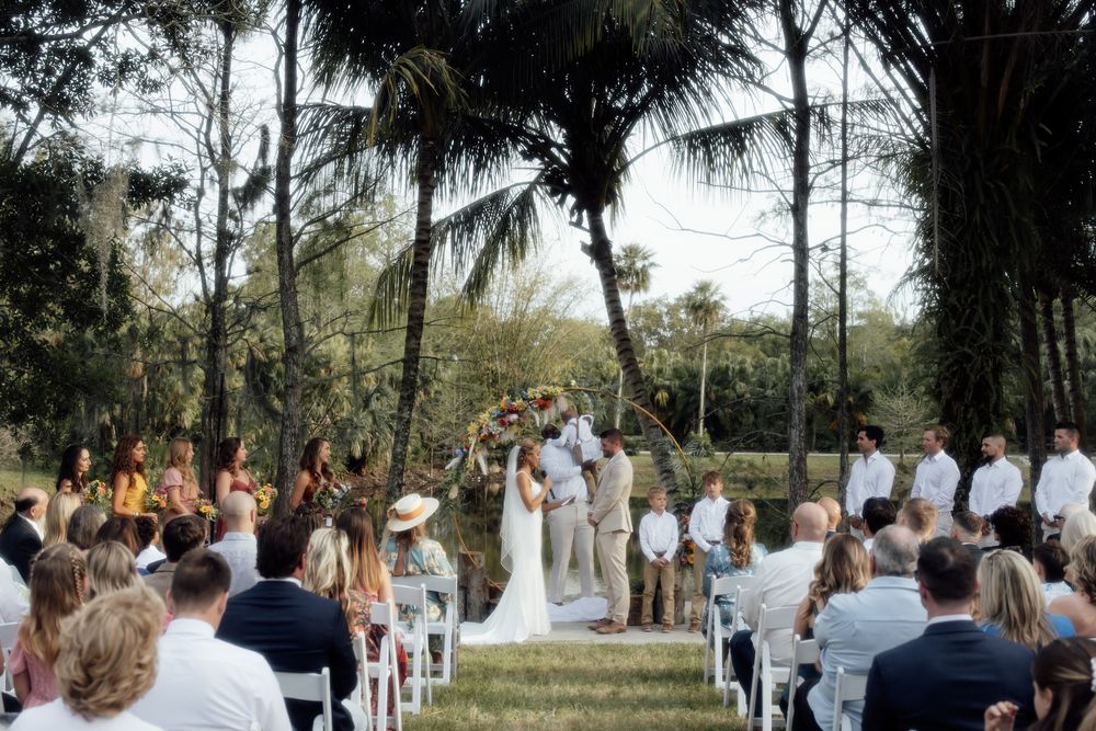 Panoramic view of an outdoor wedding ceremony under palm trees with guests seated in white chairs by the water.