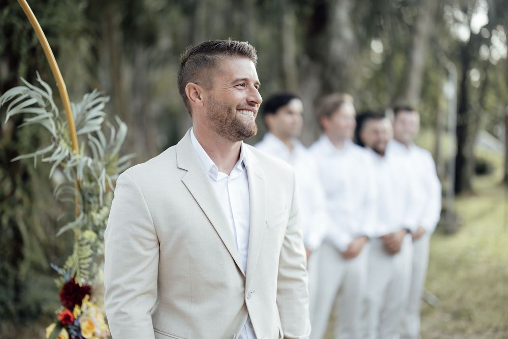 Wedding party in light colored attire standing in line under trees outdoors.