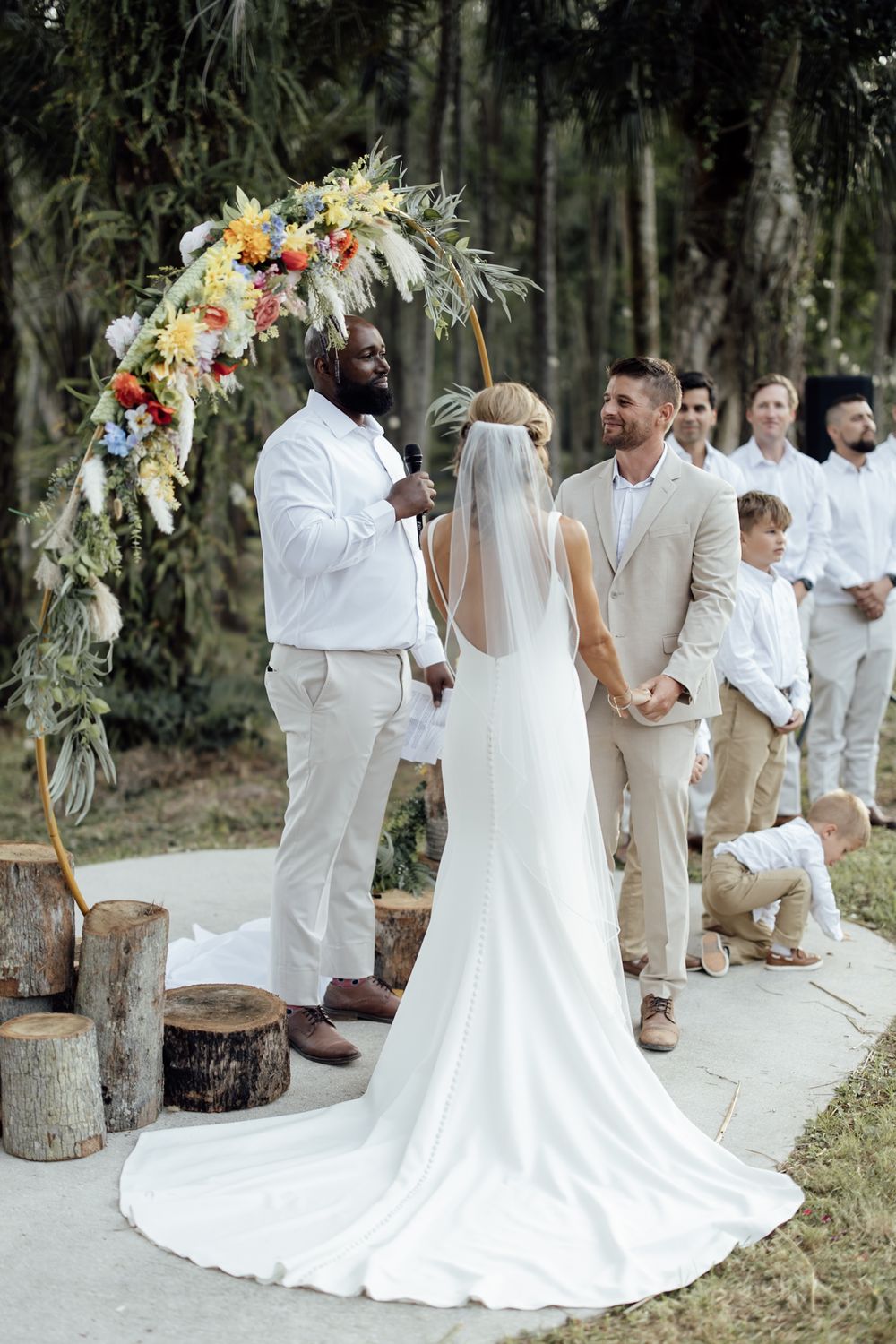 An outdoor wedding ceremony under a floral arch decorated with colorful flowers and greenery.