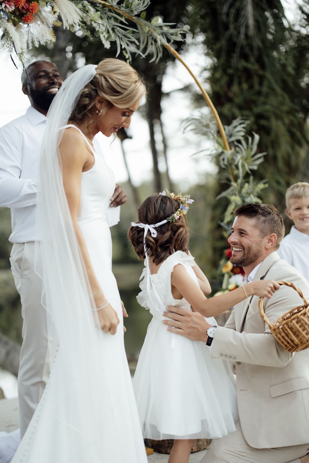 A couple shares a joyful moment during their outdoor wedding ceremony in white attire and surrounded by greenery.