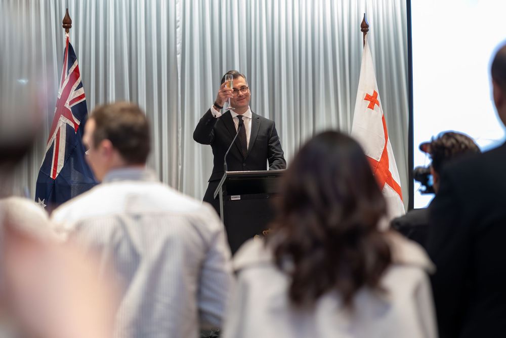 Georgian Ambassador delivering an address from the podium at a national day event, flags behind.