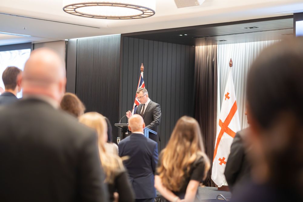 Audience listening to a speech at a diplomatic reception, stage and flags in view.