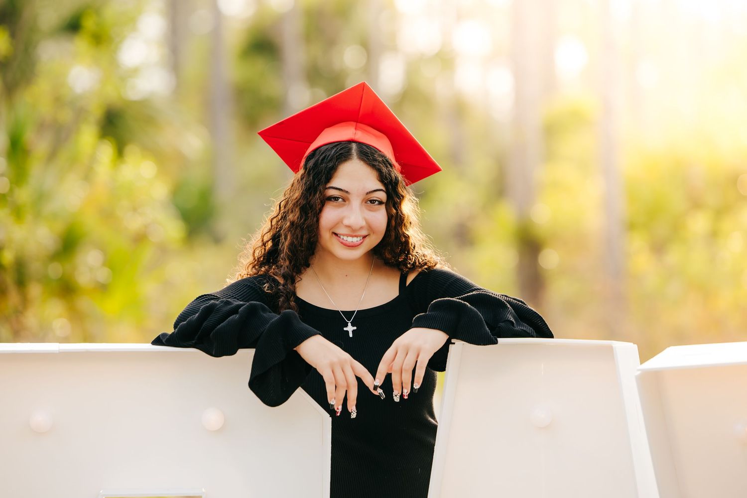 Graduate in black gown and red cap leaning on white railing with blurred natural background.