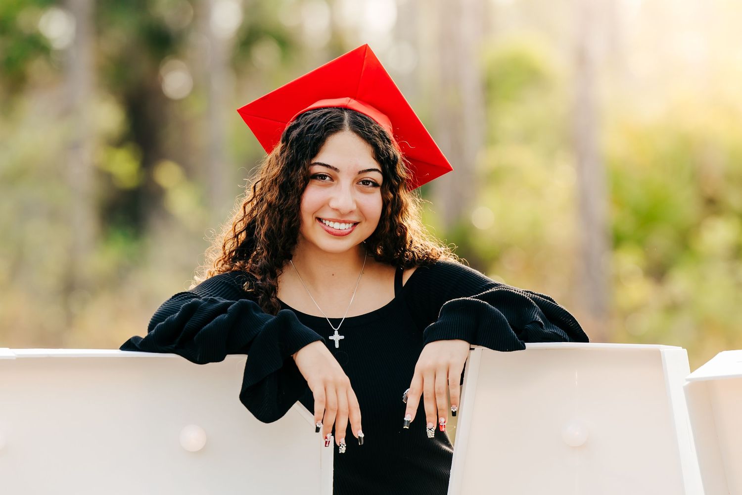 Graduate in cap and gown poses outdoors while leaning on white surface in natural setting.