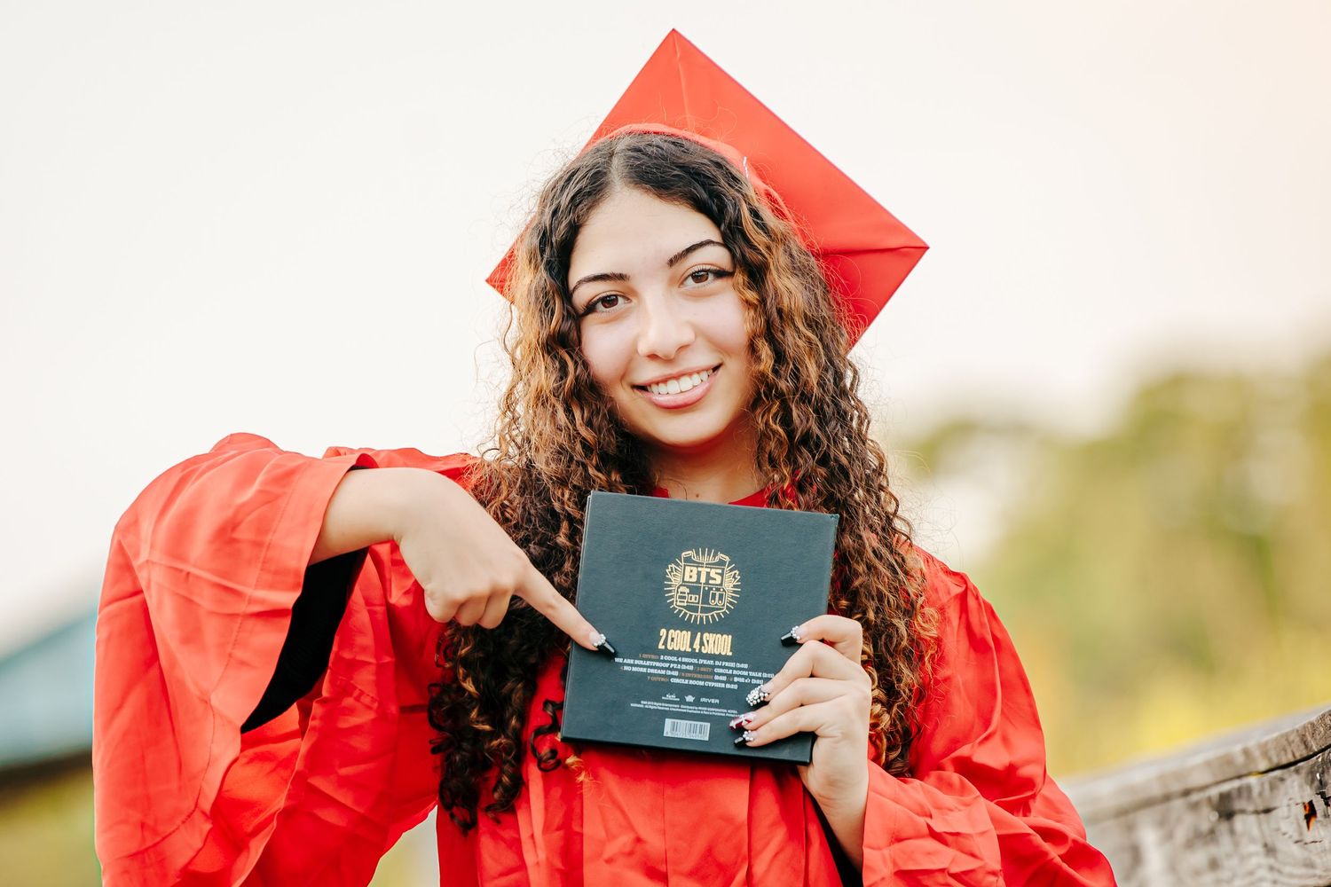 Graduate in bright red cap and gown holds up diploma or passport while celebrating outdoors.