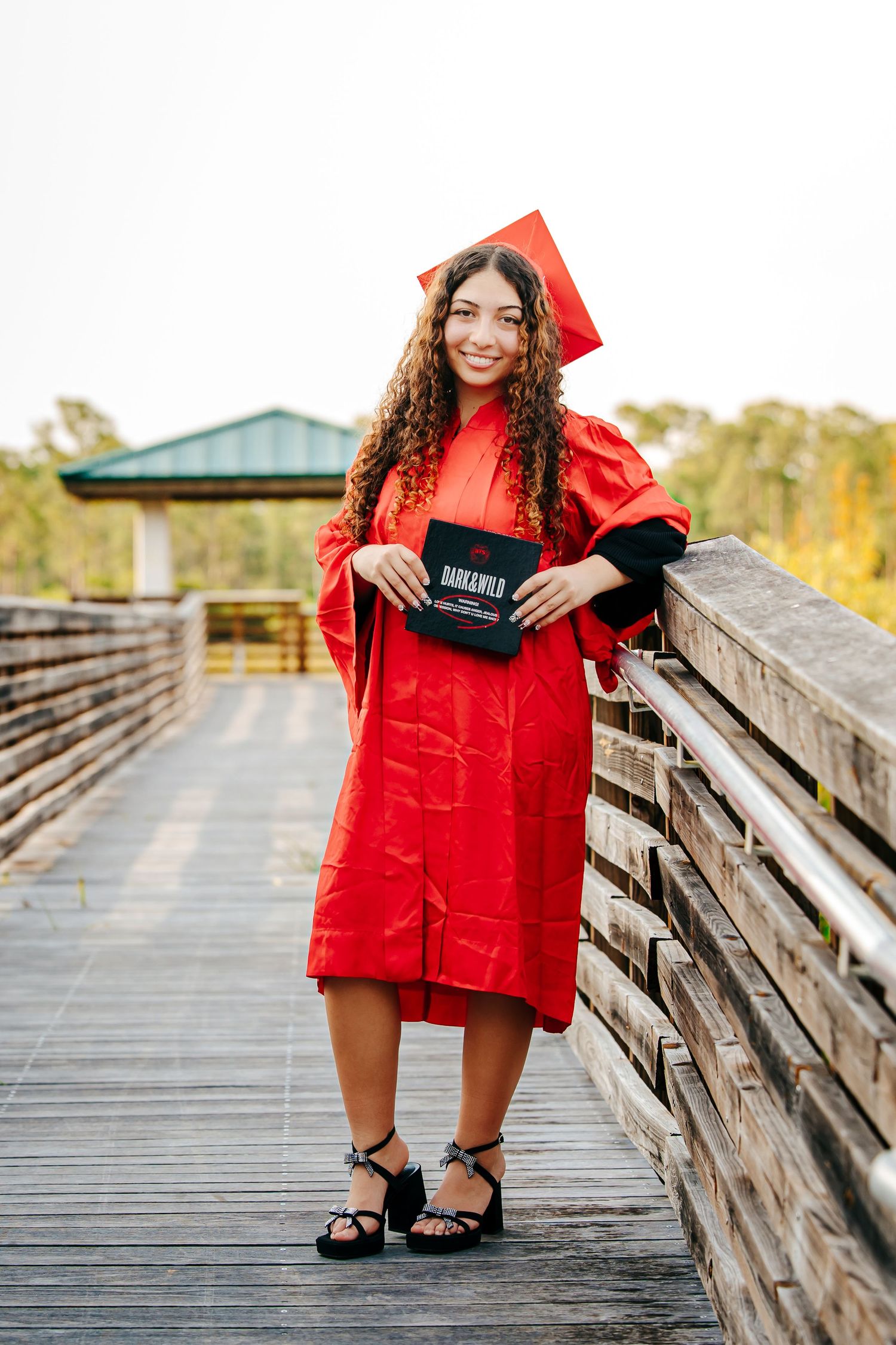 A person in a bright red graduation gown poses on a wooden boardwalk on a sunny autumn day.