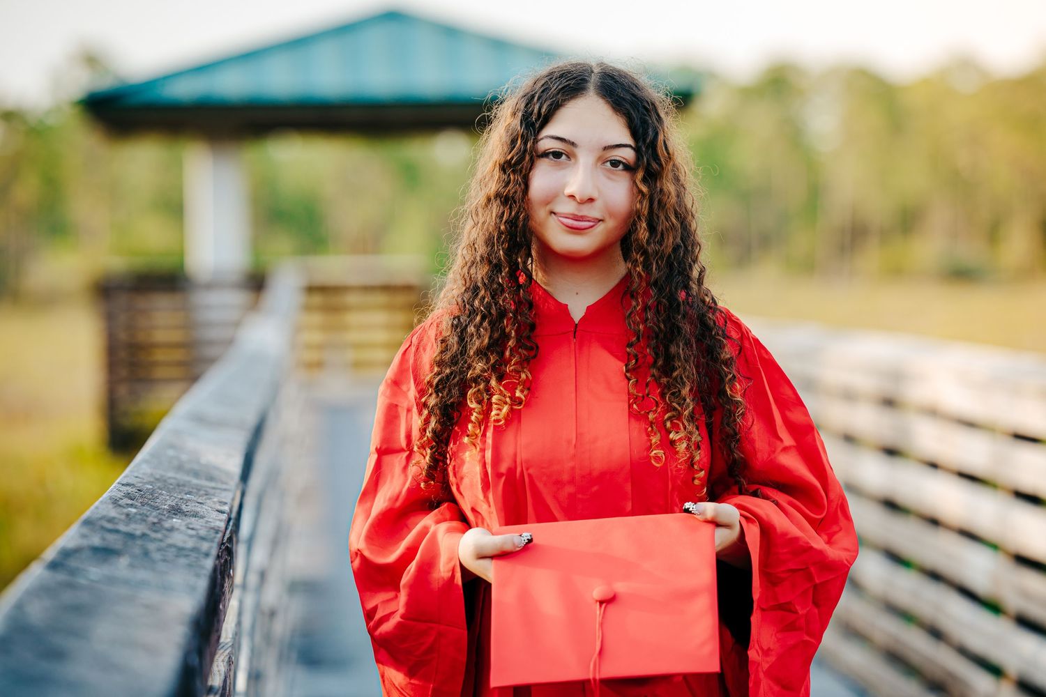 Graduate in bright red cap and gown holds matching diploma holder while standing on wooden deck outdoors.