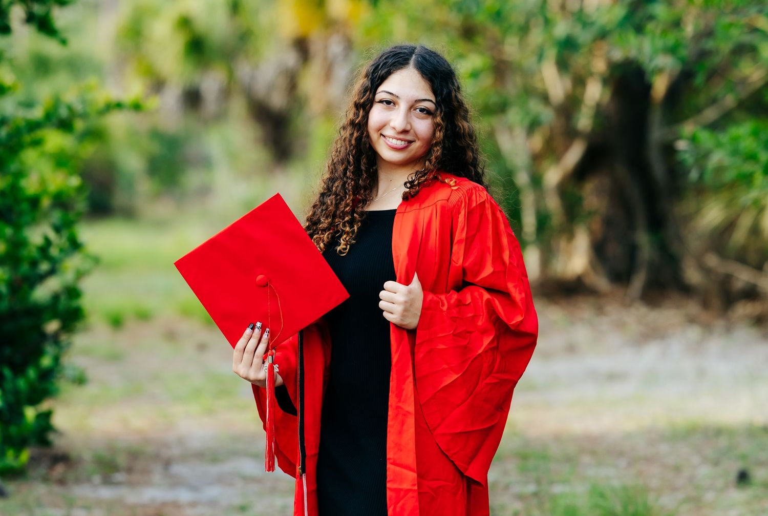 Graduate wearing red academic regalia holds red folder while posing outdoors on graduation day.
