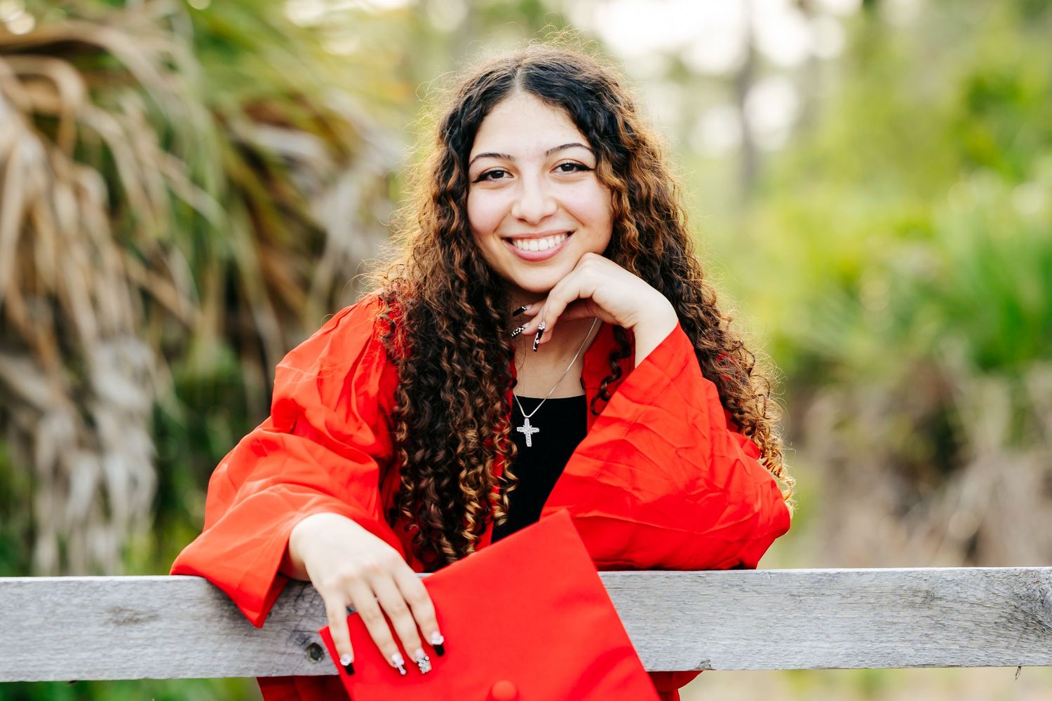 A person with curly dark hair wearing a bright red sweater leans on a wooden fence outdoors with a natural background.