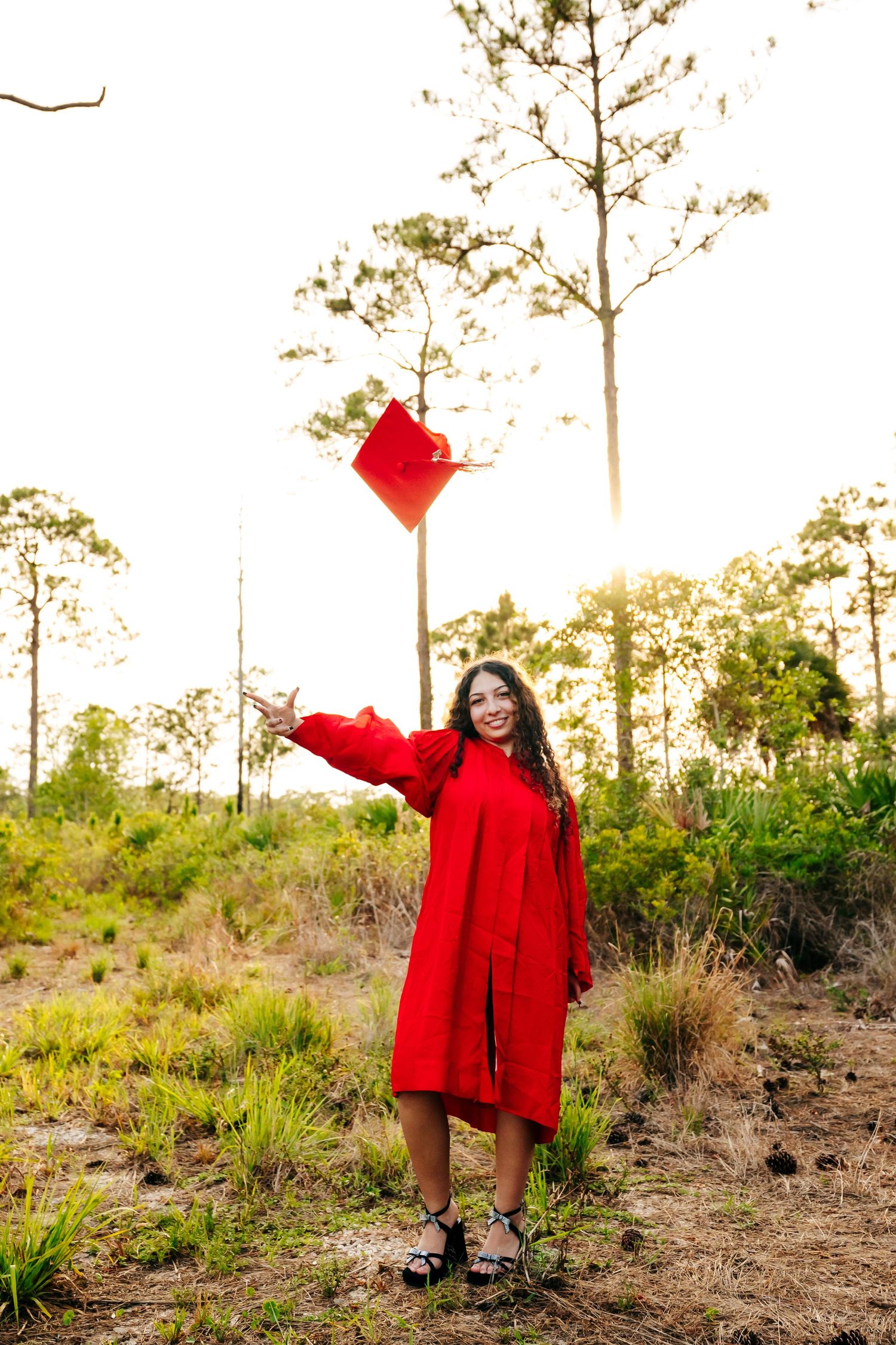 Graduate in a bright red gown holds their cap outdoors against a scenic backdrop of tall trees at sunset.