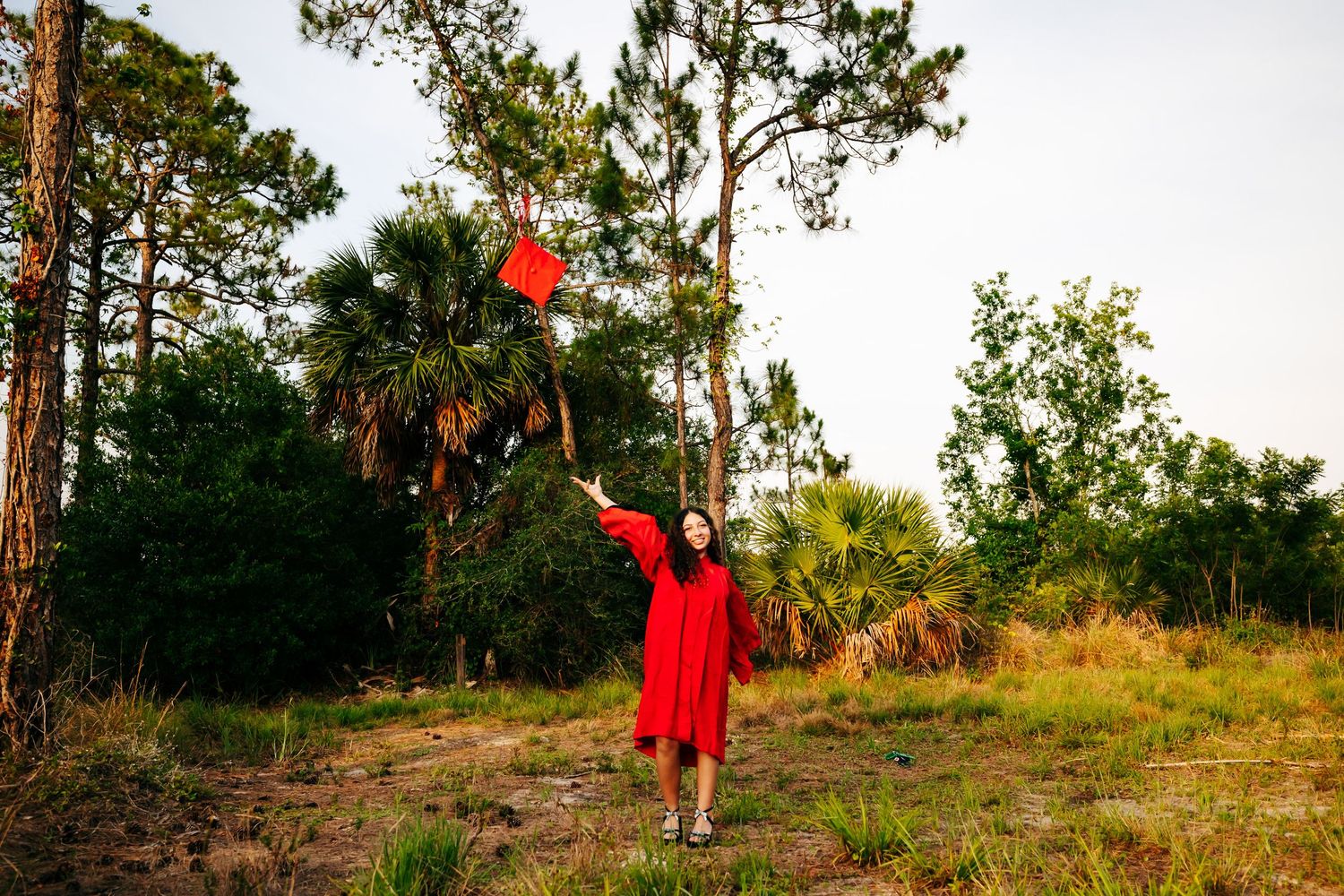 A red dress stands out against a natural green landscape with tall pine trees along a winding path at sunset.