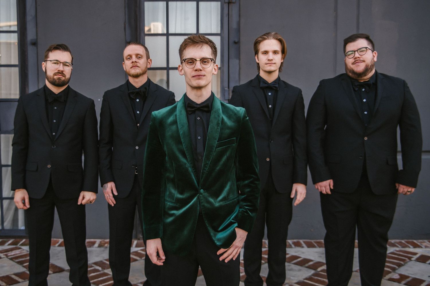Groomsmen in black suits and dark green velvet jackets pose together for a formal wedding party photo.