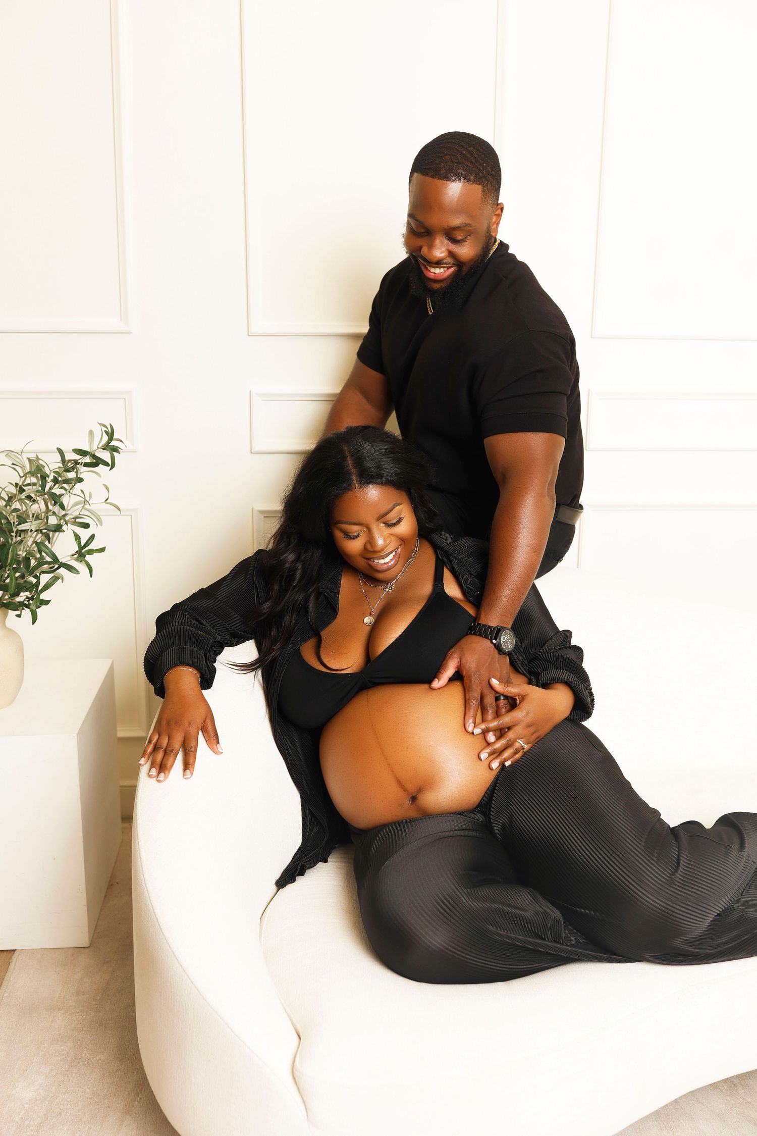A couple sits intimately together on a white couch for their maternity photo session.