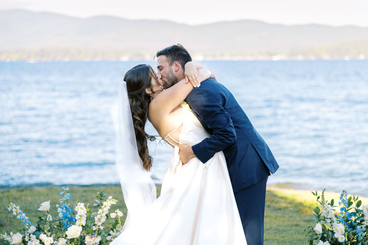 A couple shares a romantic embrace during a scenic outdoor wedding photoshoot by a lake with mountains in the distance.