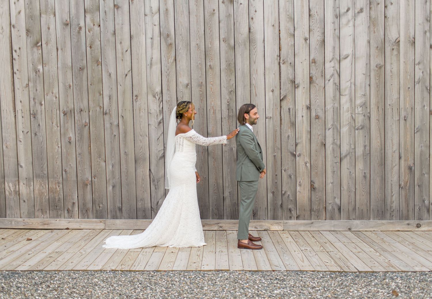 A bride and groom share a playful moment standing back to back against a wooden wall in a first look photo.