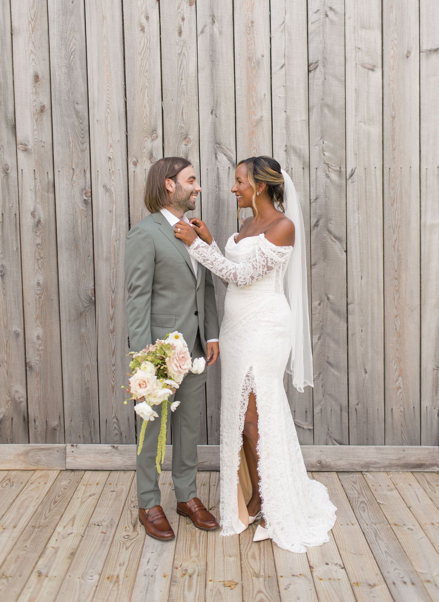 Romantic wedding couple shares tender moment against rustic wood wall backdrop with bride holding white floral bouquet.