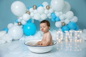 Adorable baby's first birthday setup with blue and white balloons, glowing ONE sign, and bathtub photoshoot props.