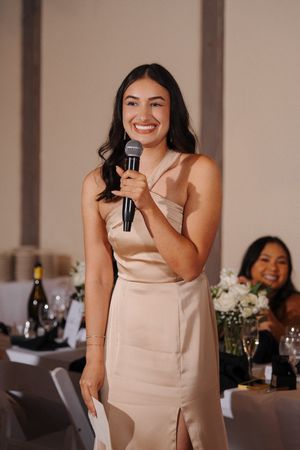 A bridesmaid in a champagne satin dress gives a speech at a wedding reception with flowers and wine bottles visible.
