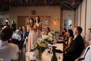 Wedding reception guests gather at long tables while a bridesmaid gives a toast at a rustic indoor venue.