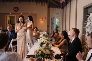 Wedding guests watch as a speaker in a champagne-colored dress gives a toast at an elegant reception decorated with flowers.