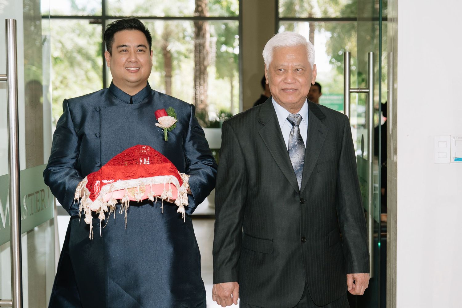 Groom in navy ao dai carrying red lacquered wedding gift box walking with elderly man at ceremony.