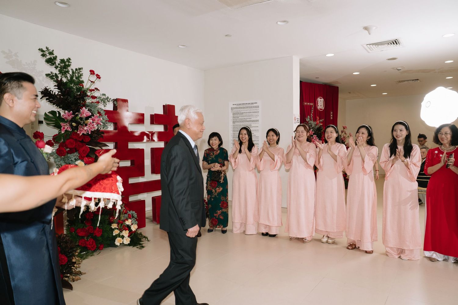Elderly man greeted by bridesmaids in pink ao dai dresses at a traditional Vietnamese wedding ceremony.