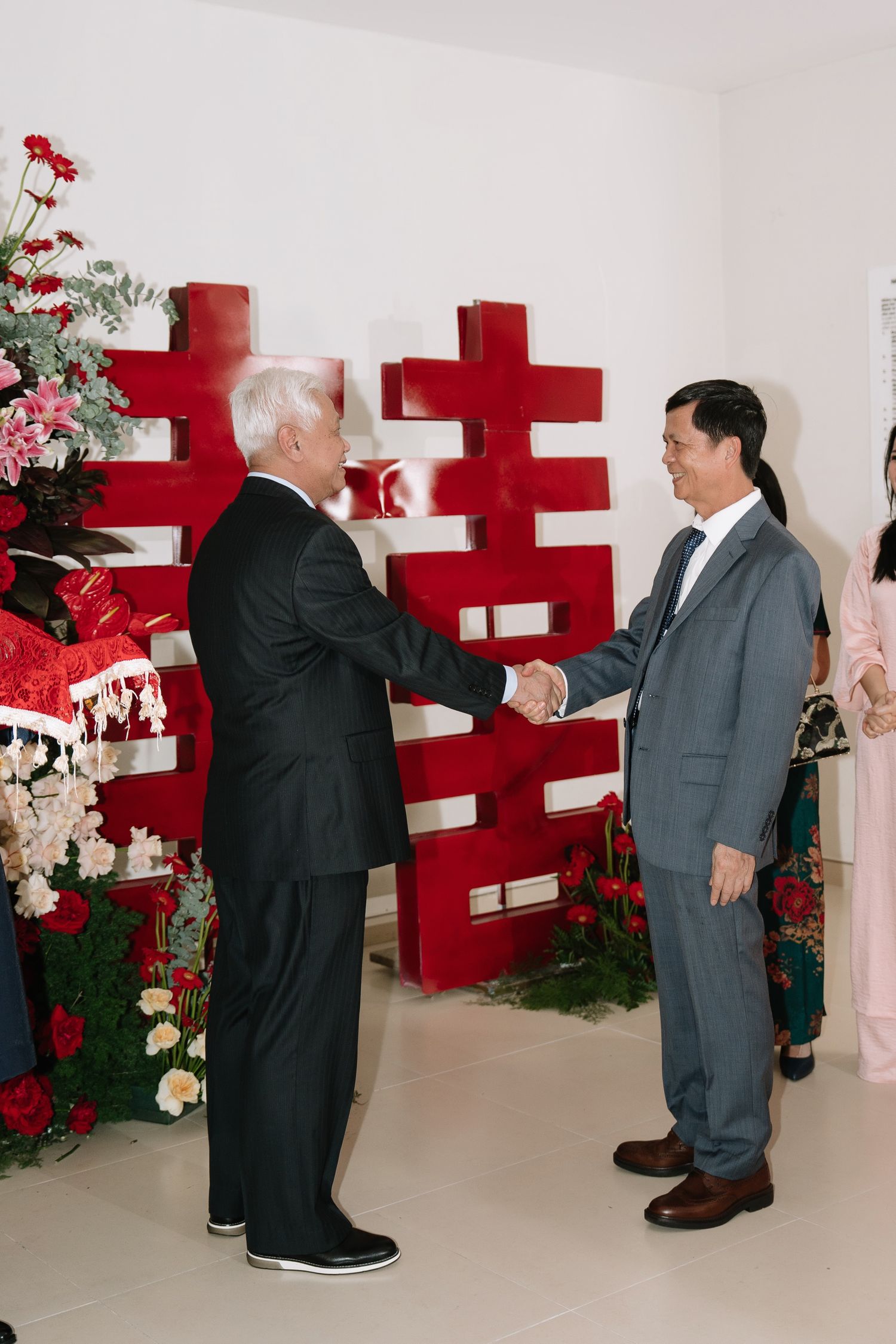 Two elderly men in formal suits shaking hands before large red double happiness symbol at wedding.