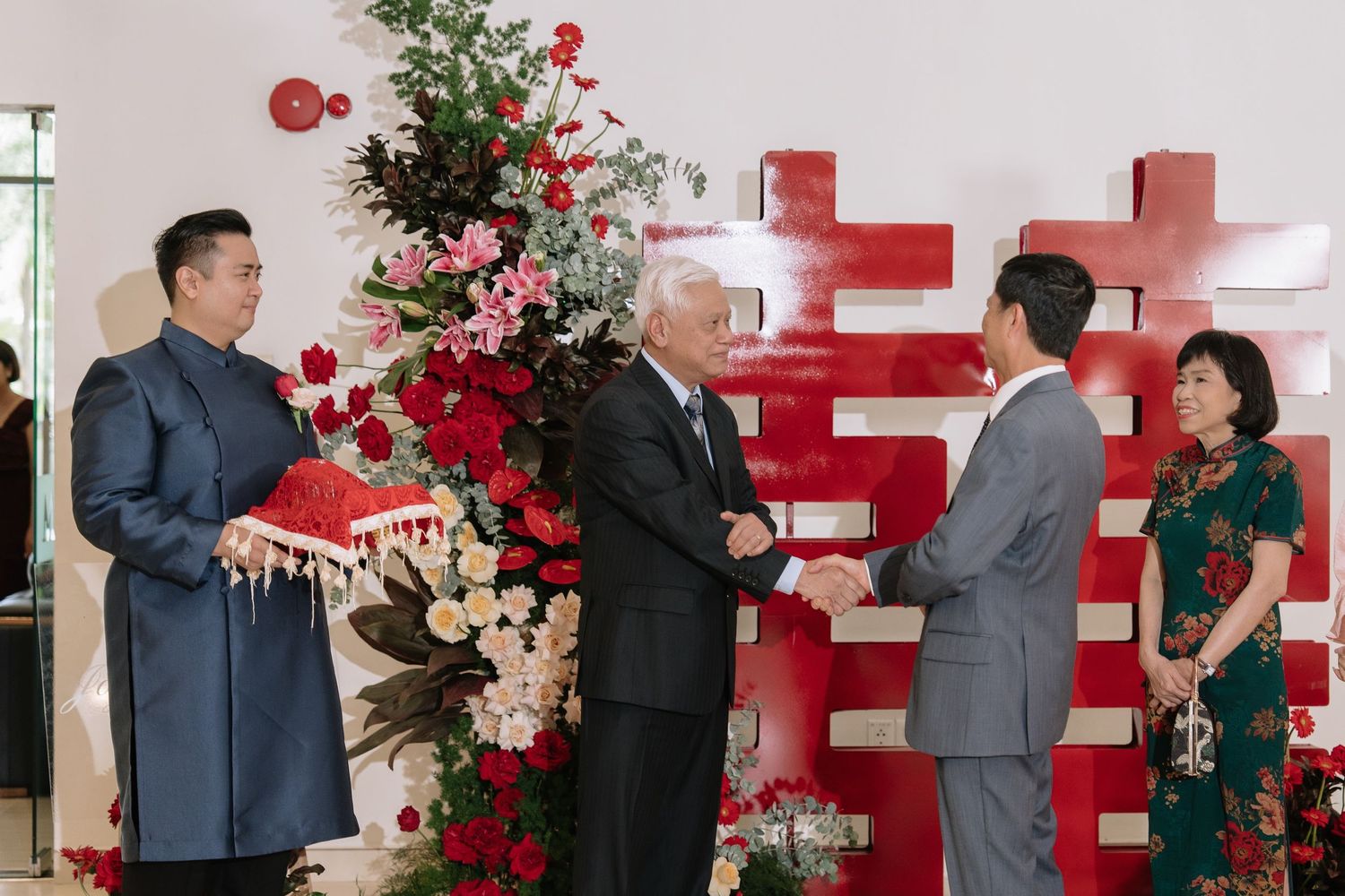 Groom presenting red gift box as two older men shake hands beside floral arrangement at Vietnamese wedding.