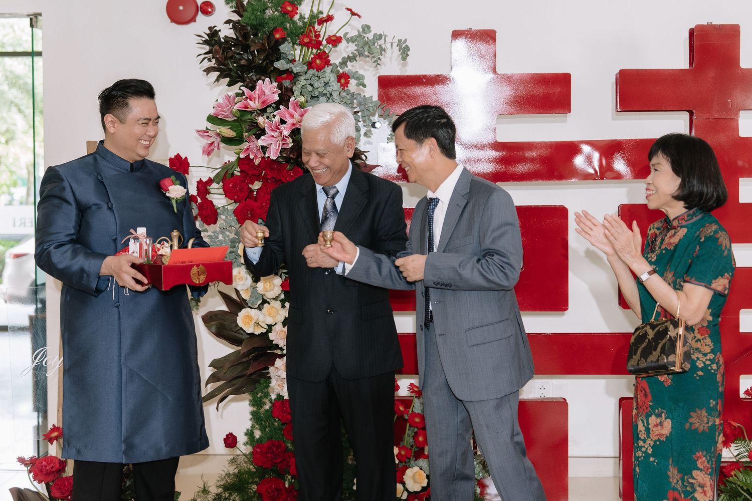 Groom and family exchanging red gift boxes and toasting beside floral decor at traditional Vietnamese wedding.