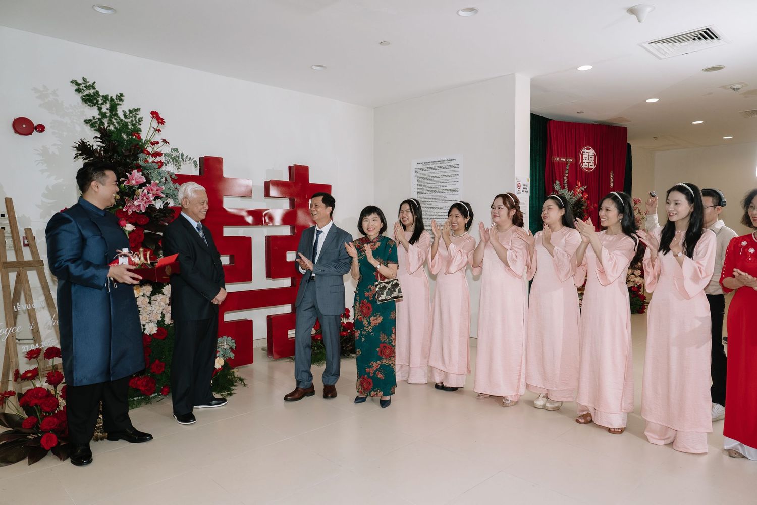 Wedding party in pink ao dai dresses applauding couple and elders beside red double happiness decor.