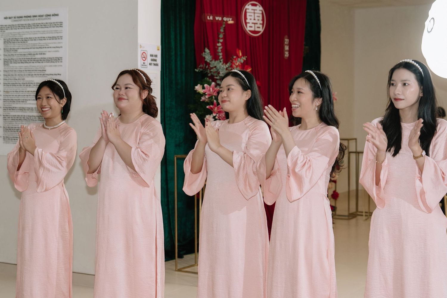 Five women in matching pink ao dai dresses clapping joyfully at a traditional Vietnamese wedding ceremony.