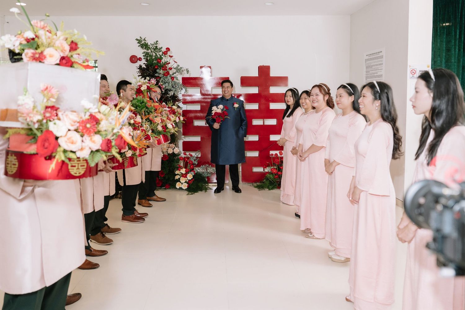Vietnamese wedding ceremony with bridesmaids in pink ao dai and groomsmen presenting floral gifts indoors.