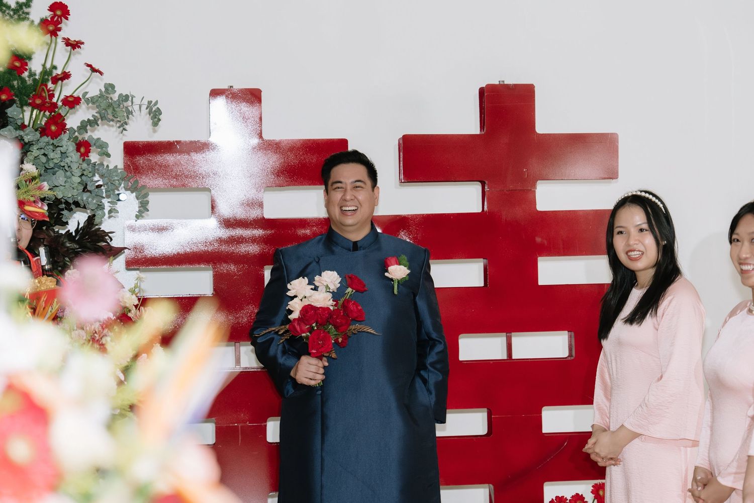 Groom in navy ao dai holding red roses stands before red double happiness wedding backdrop.