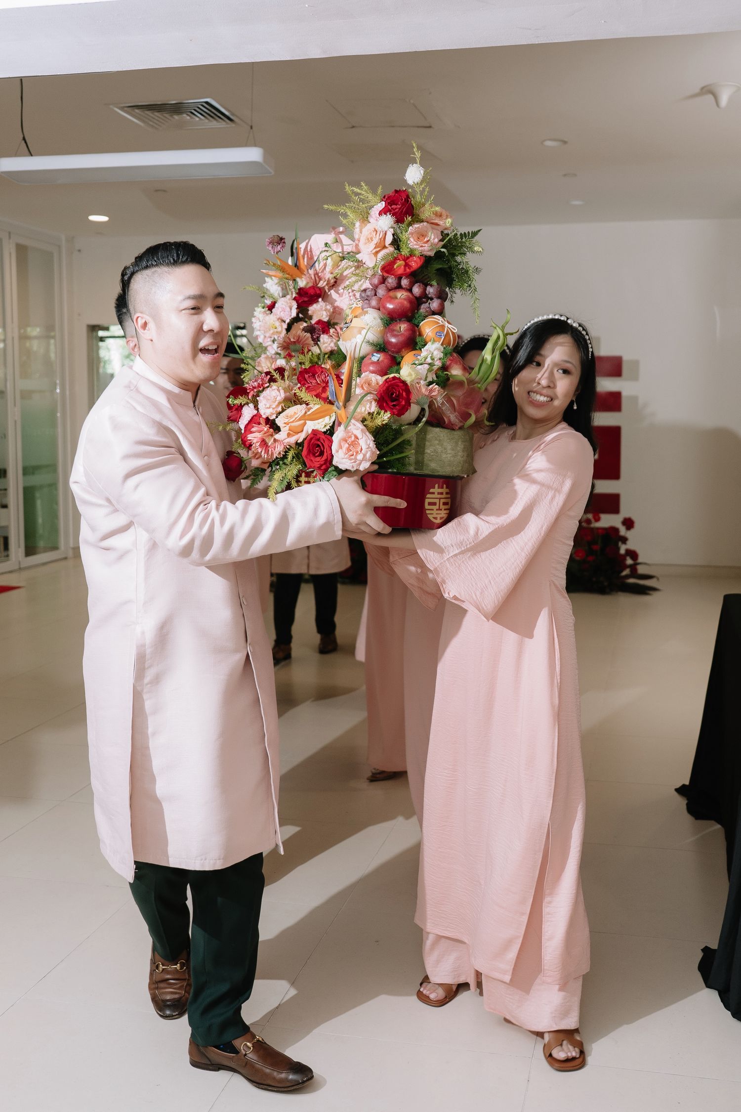 Man and woman in pink ao dai exchange large floral arrangement during Vietnamese wedding ceremony.