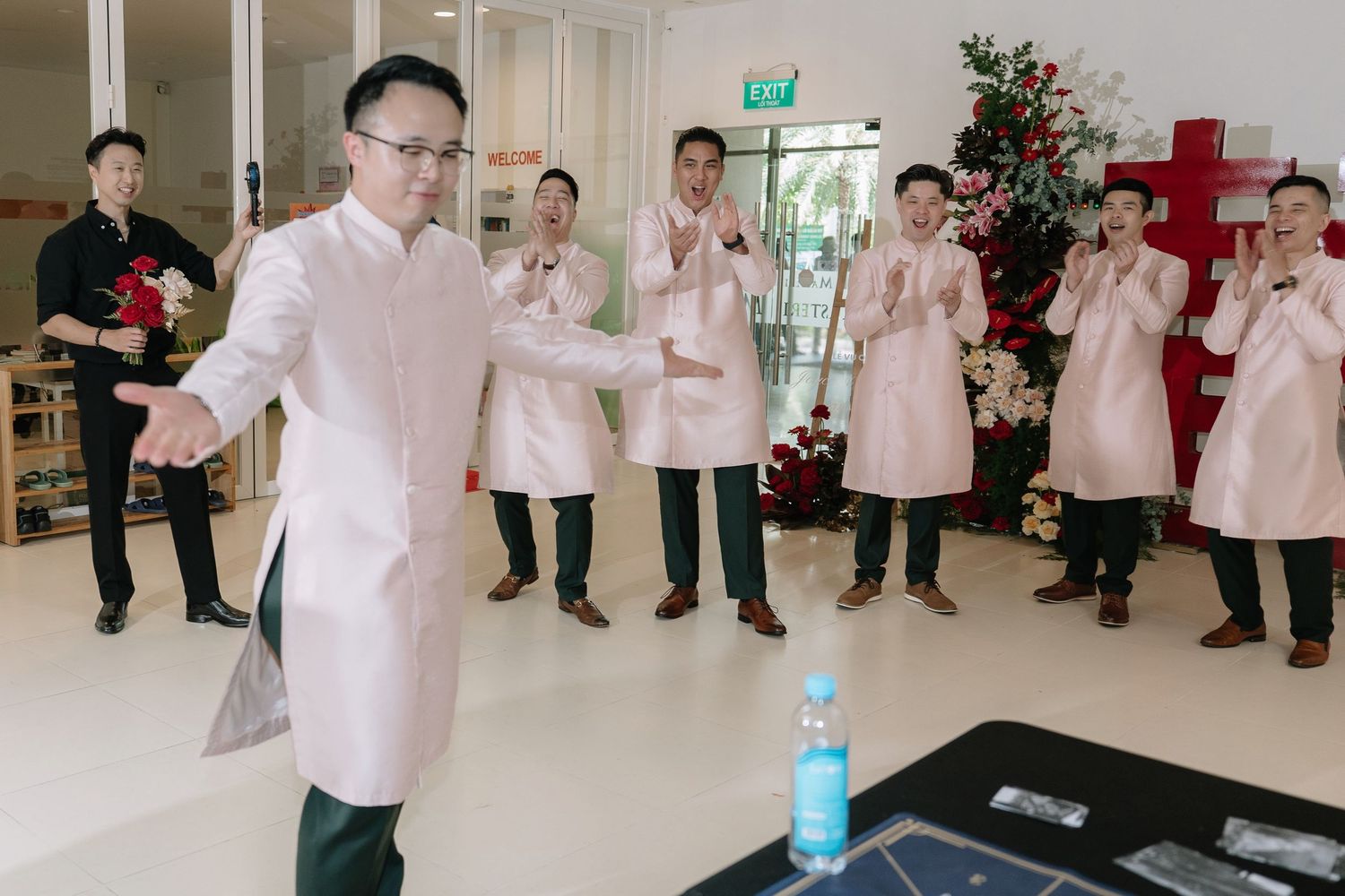 Groomsmen in pink ao dai clap and dance during fun Vietnamese wedding celebration indoors.