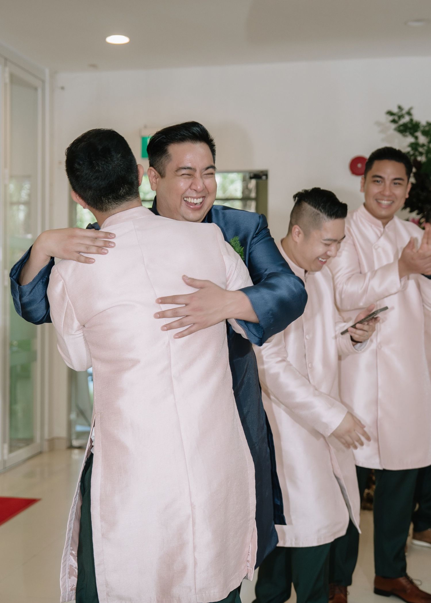 Groom in blue ao dai shares warm embrace with groomsman at Vietnamese wedding celebration.