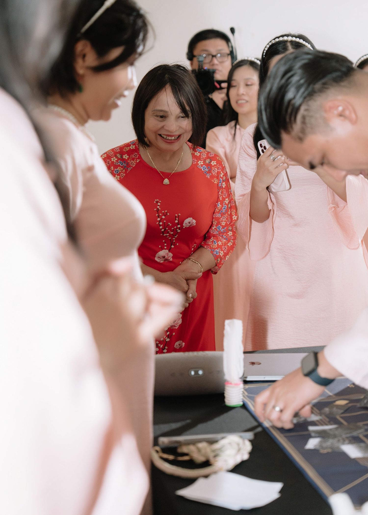 Older woman in red ao dai smiles warmly surrounded by bridesmaids in pink at a Vietnamese wedding ceremony.