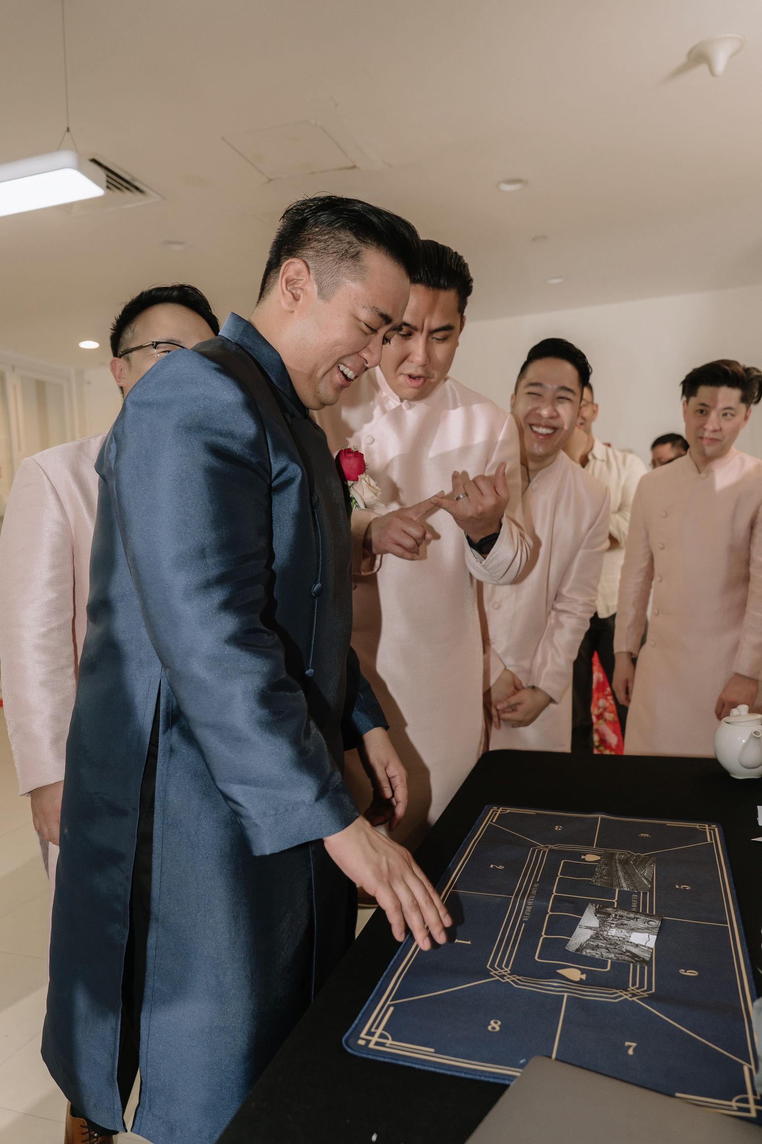 Groom in navy ao dai laughs while pointing at wedding game board surrounded by smiling groomsmen in pink.