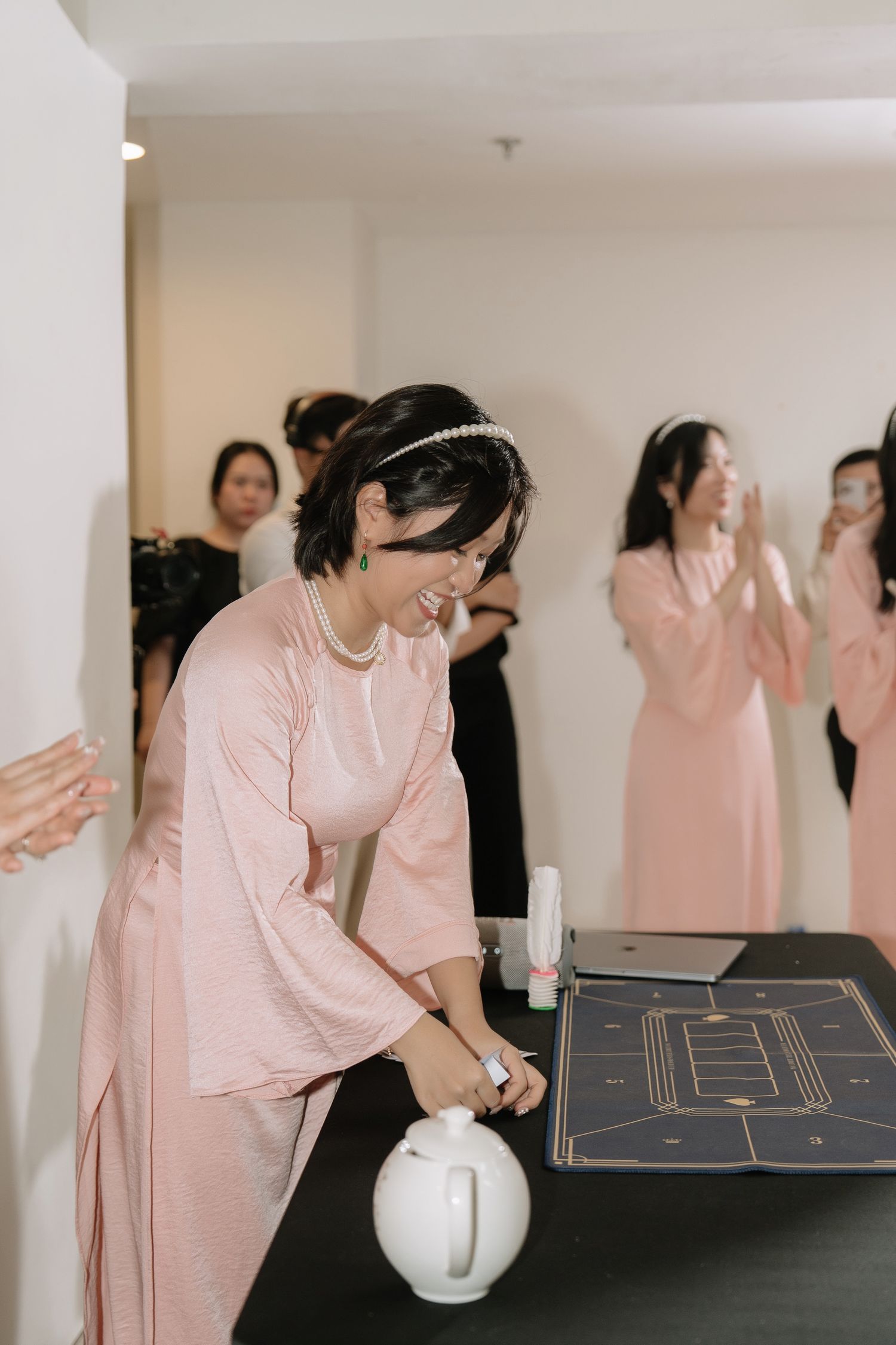 Bridesmaid in pink ao dai with pearl headband smiles while interacting with wedding game board at ceremony.