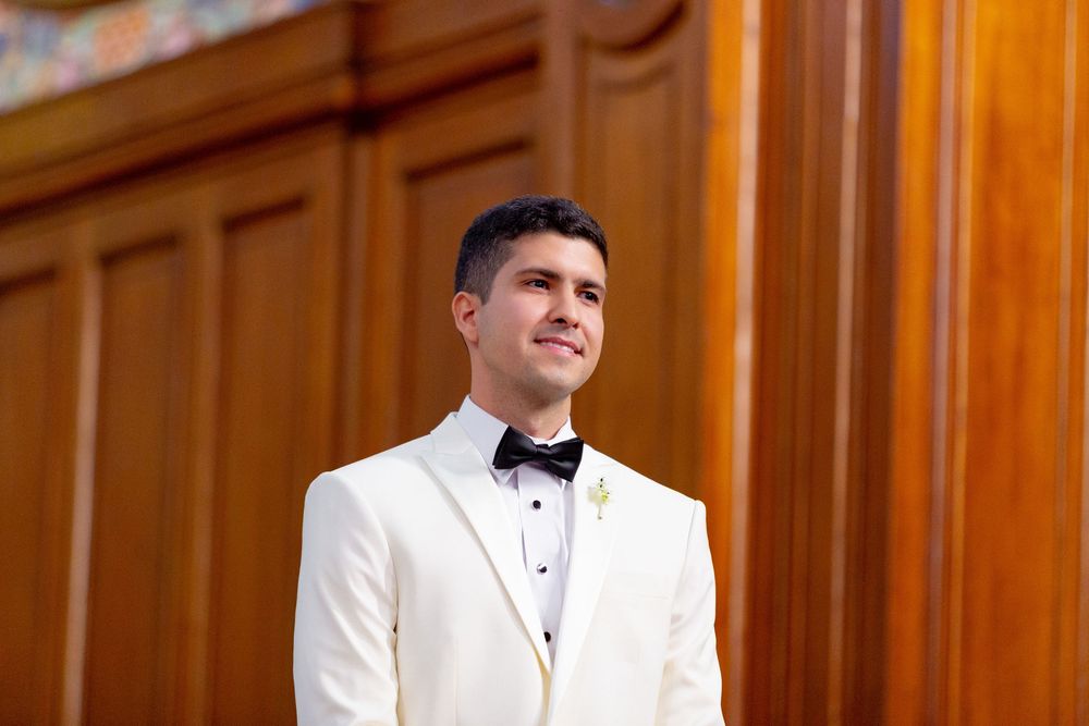 A person in a white tuxedo jacket and black bow tie poses casually against a wooden panel backdrop.