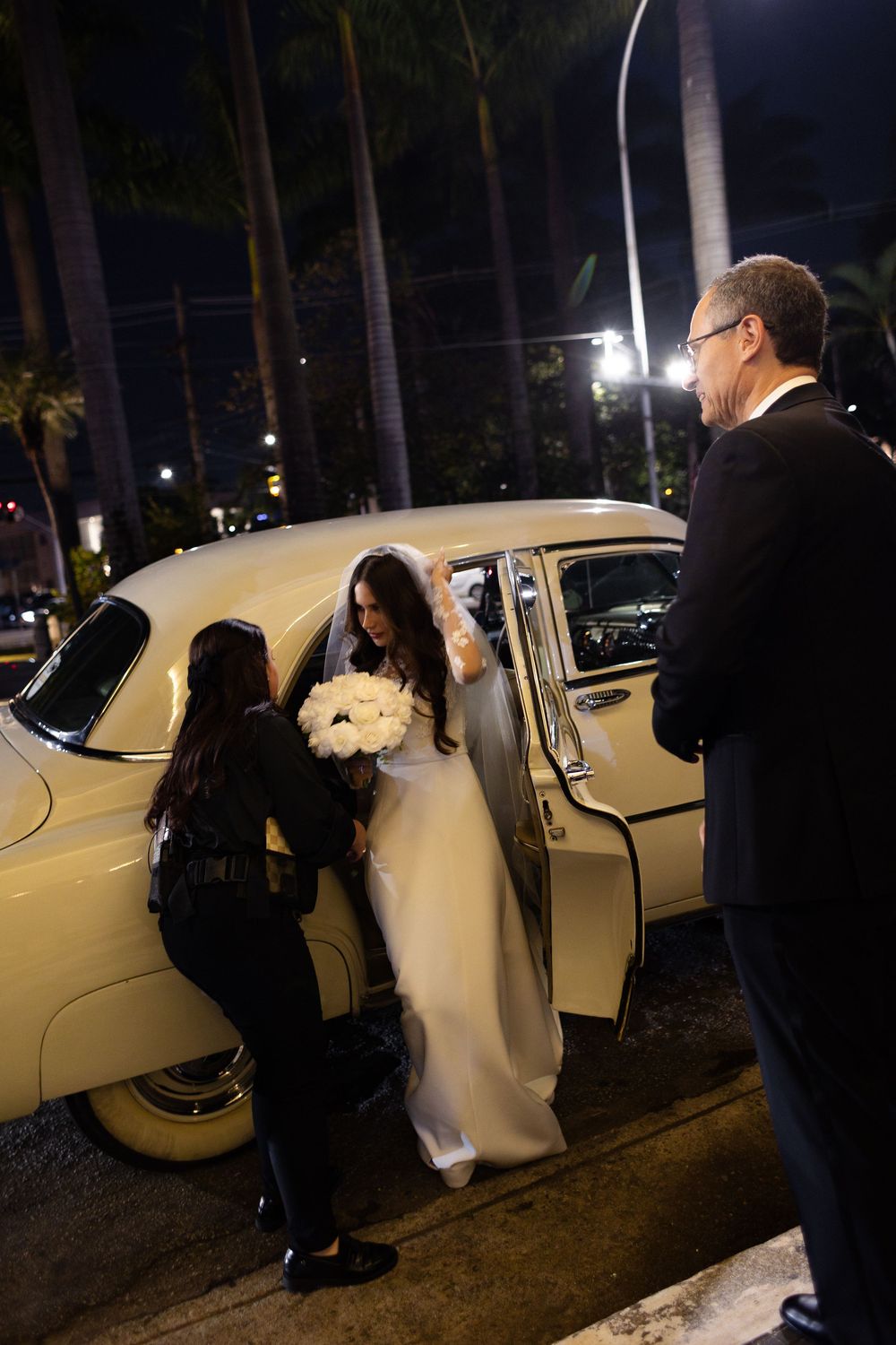 At night, someone opens a car door for a small white dog to enter a vintage cream-colored vehicle.