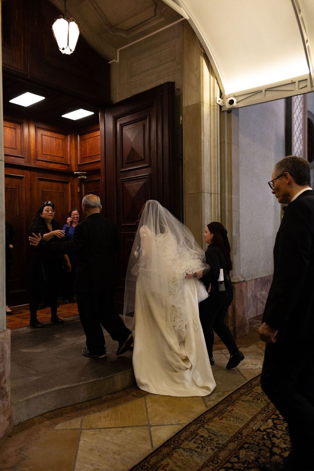 A wedding party prepares and positions themselves in an elegant hallway with wood paneling and ornate carpeting.