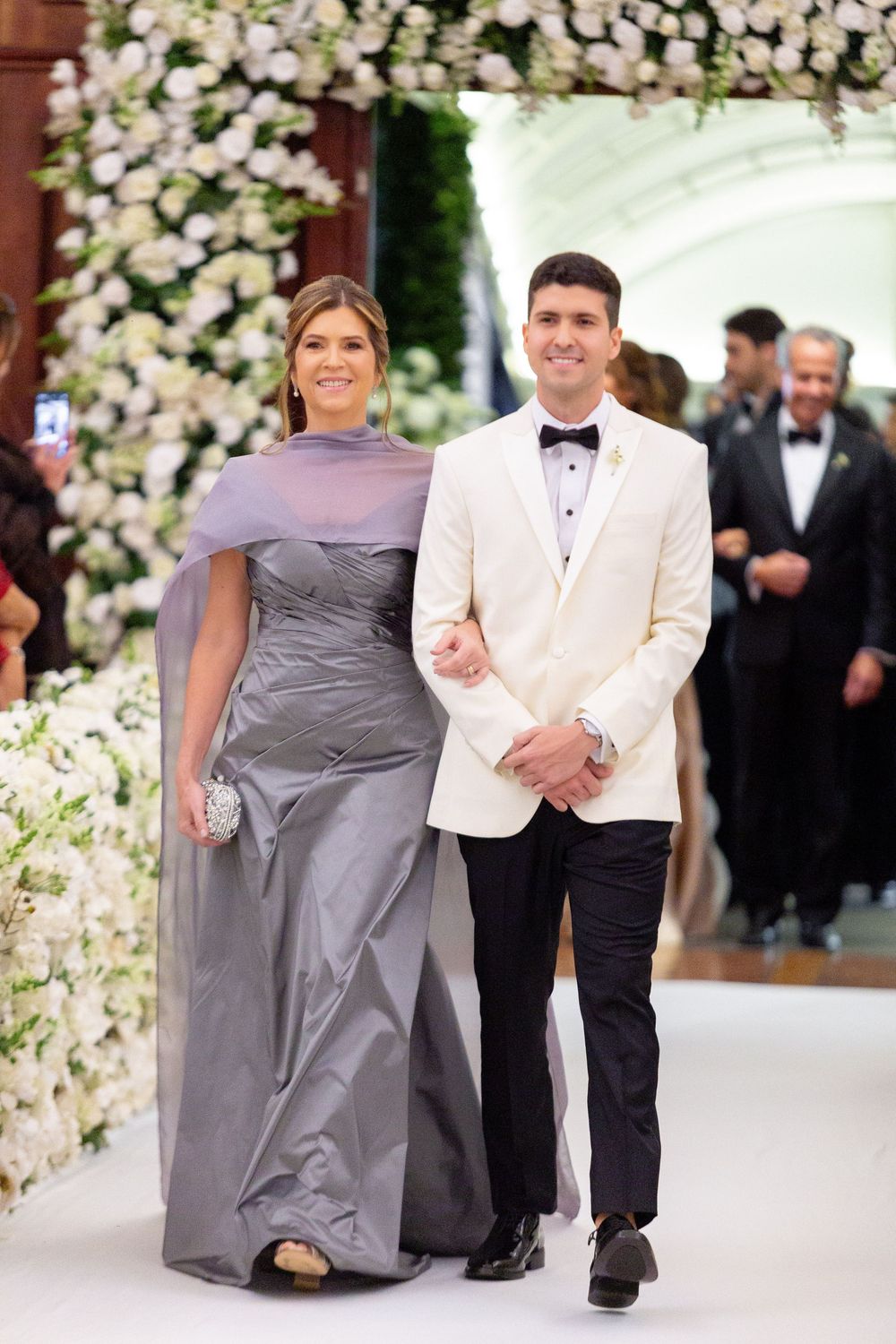 Wedding party members in grey dresses and white tuxedos walk down an aisle decorated with white floral arrangements.