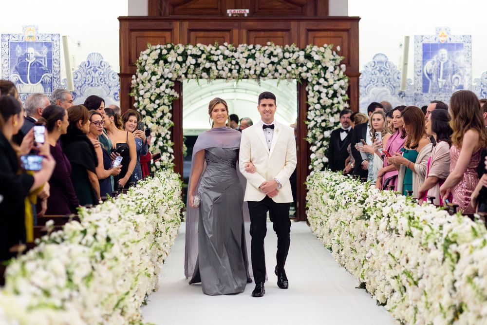 A wedding ceremony with a mother and groom walking down an aisle decorated with white floral arrangements and archway.